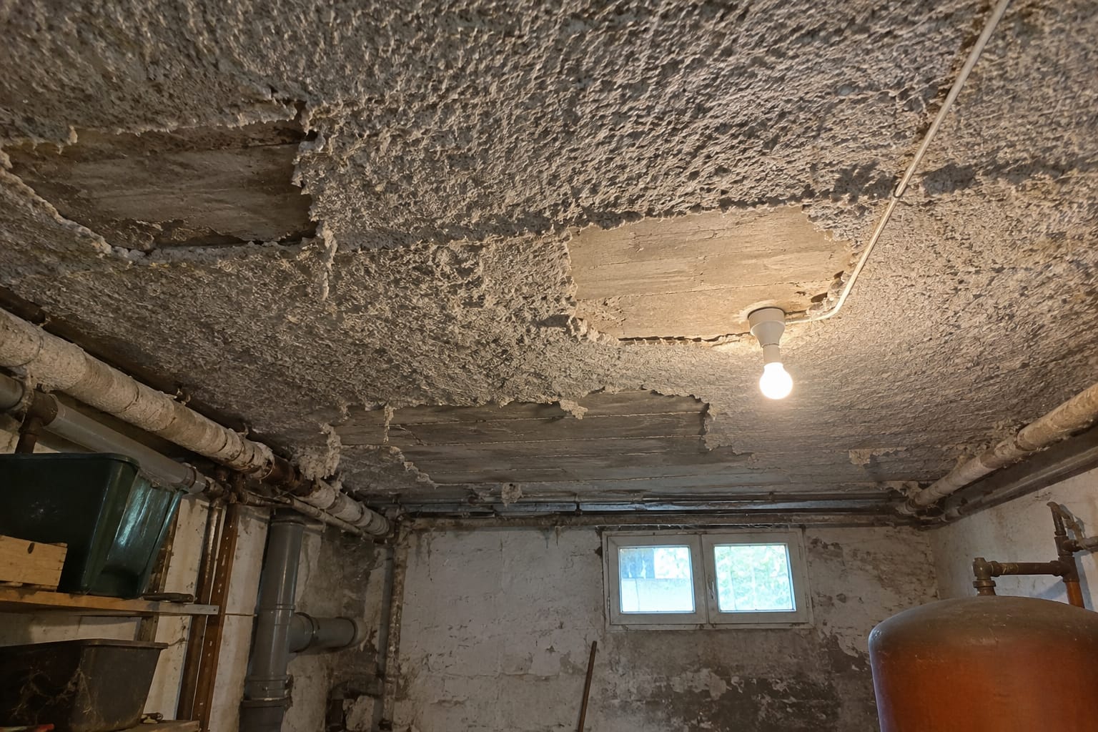 Plafond de sous-sol couvert d’un enduit fibreux gris projeté, décollé par plaques, des fragments tombés au sol.