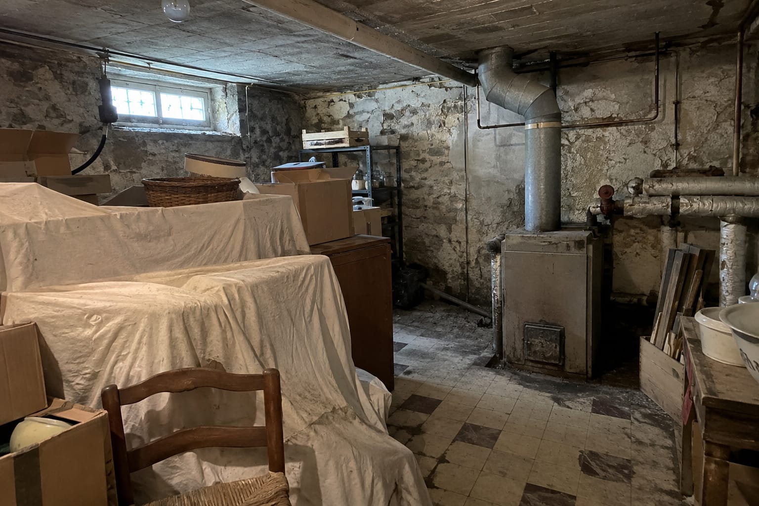 Un sous-sol encombré de cartons et de meubles bâchés, derrière lesquels un vieux conduit gris monte au plafond.