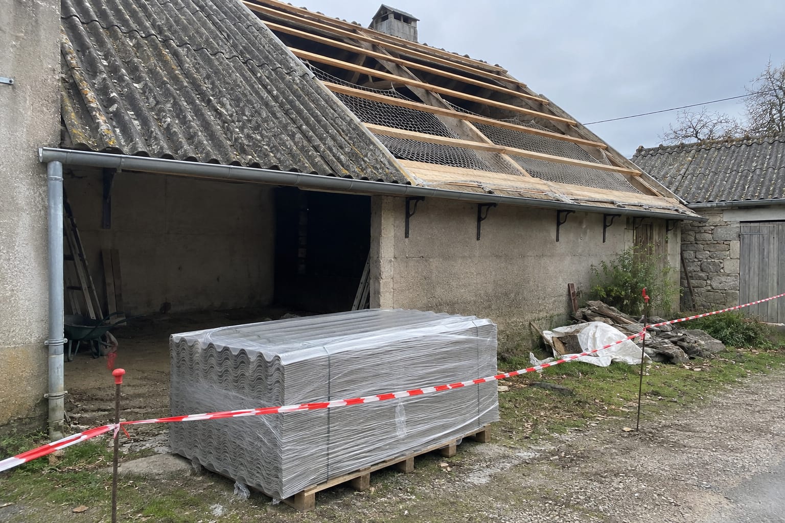 Une pile de plaques ondulées grises entières, filmée sur une palette au pied d’un bâtiment agricole à moitié découvert, un filet tendu sous les pannes.